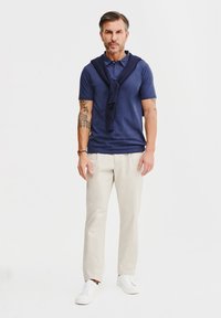 Granatowa koszulka polo z krótkim rękawem i kołnierzykiem, noszona z jasnobeżowymi chinosami i białymi trampkami. Granatowy sweter jest przewiązany na ramionach.