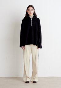 Schwarzer übergroßer Pullover mit hohem Kragen und silbernem Reißverschluss, kombiniert mit cremefarbenen weit geschnittenen Hosen mit seitlichen Streifen, getragen mit dunklen Schuhen.