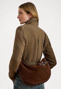 Borse a spalla in suede marrone con forma curva e accenti in pelle. Presenta una fibbia argentata e una superficie liscia, indossata a tracolla.