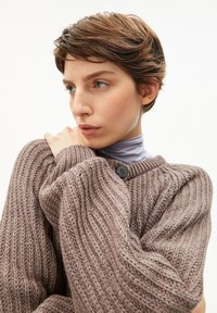 Junge Person mit kurzen braunen Haaren, die einen übergroßen, grob gestrickten taupefarbenen Pullover trägt, hält den Pullover mit der linken Hand nah an sich und schaut nach links.
