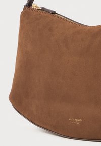 Brown suede handbag featuring a curved shape, gold hardware, and embossed logo. Soft texture with a smooth zipper closure.