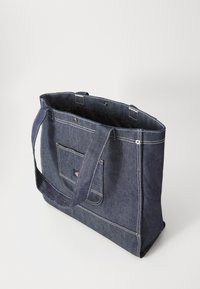 Denimväska med ett stort huvudfack, slitstarka remmar, vit sömnad, framficka och en liten logodetalj nära botten.