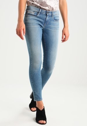 Frau trägt hellblaue Skinny-Jeans und schwarze offene Sandaletten, steht mit einem Bein leicht vor das andere gekreuzt.