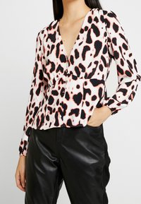 Blusa de estampado de leopardo en tejido ligero, con un escote en V profundo, mangas fruncidas y cintura ajustada con botones. Combinada con pantalones negros.