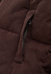 Close-up of a partially open silver zipper on dark brown corduroy fabric with visible ridged texture and padded stitching lines.