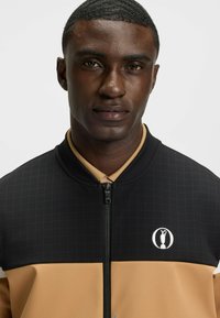 Black and tan zip-up jacket with a grid pattern, featuring a round logo and a solid tan lower section; textured fabric, ribbed collar.