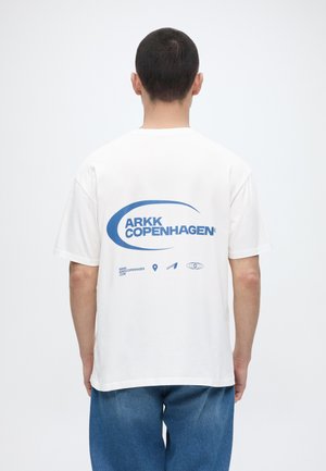 Mann som har på seg hvit T-skjorte med blå "ARKK Copenhagen"-logo og ikoner på baksiden, kombinert med blå jeans, med ryggen til på hvit bakgrunn.