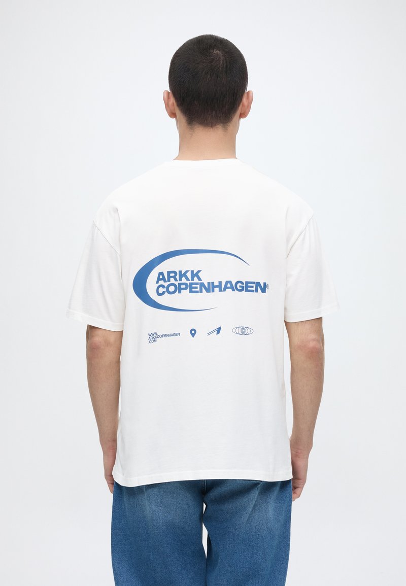 Άνδρας που φοράει άσπρο T-shirt με μπλε λογότυπο και εικονίδια "ARKK Copenhagen" στην πλάτη, συνδυασμένο με μπλε τζιν, γυρισμένος πάνω σε λευκό φόντο.