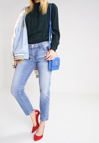 Svetle modrá bomber bunda se záplatami, tmavě zelená blůza, světle modré džíny, červené vysoké podpatky a malá modrá kabelka přes rameno.