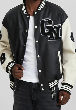 Giacca varsity nera e crema con corpo in pelle, polsini a costine e toppe ricamate. Dotata di bottoni a pressione e dettaglio portachiavi.