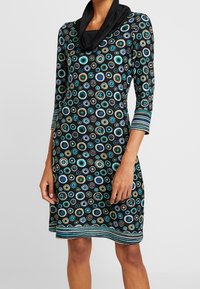 Czarna sukienka z dekoltem w kształcie kaptura, z kolorowymi koncentrycznymi okręgami oraz paskowanymi akcentami na rękawach i u dołu. Miękka tkanina, długość do kolan.