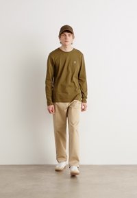 Olivgrünes Langarmhemd mit kleinem Logo, beigen Hosen, weißen Turnschuhen und passender Mütze. Einfaches Design vor einem hellen Hintergrund.