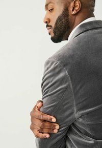 Grauvelours-Blazer mit strukturierter Oberfläche und taillierter Form. Verfügt über einen schmalen Revers und schmal geschnittene Ärmel. Die Hand ruht auf dem Arm.