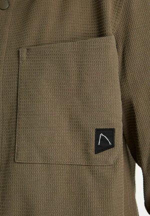 Hemd van olijfgroen stof met wafeltextuur, voorzien van een borstzak en een zwarte patch met een wit geometrisch ontwerp. Knopen aan de kraag.