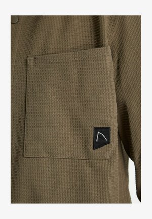 Hemd van olijfgroen stof met wafeltextuur, voorzien van een borstzak en een zwarte patch met een wit geometrisch ontwerp. Knopen aan de kraag.