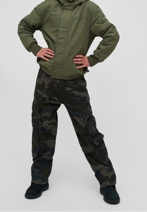 Dziecko w zielonej kurtce i czarnych spodniach bojówkach w kamuflażu stoi z rękami na biodrach na jednolitym tle.