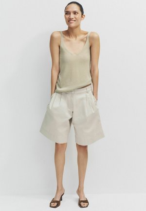 Frau lächelt, trägt ein ärmelloses beiges Oberteil, lockere, helle beige knielange Shorts mit Taschen und braune Sandalen mit offenem Zehenbereich und Absatz, steht vor einer weißen Wand.