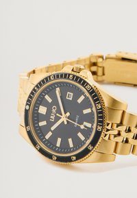 Ρολόι quartz με μαύρο καντράν, περίβλημα και μπρασελέ από ανοξείδωτο χάλυβα σε χρυσό τόνο. Διαθέτει φωτεινούς δείκτες ωρών και ένδειξη ημερομηνίας.