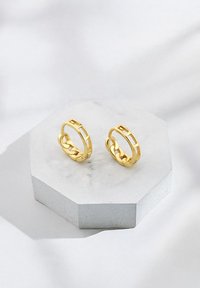 Orecchini a cerchio in tono oro con design a catena e ritagli rettangolari. Esibiti su un supporto esagonale in marmo.