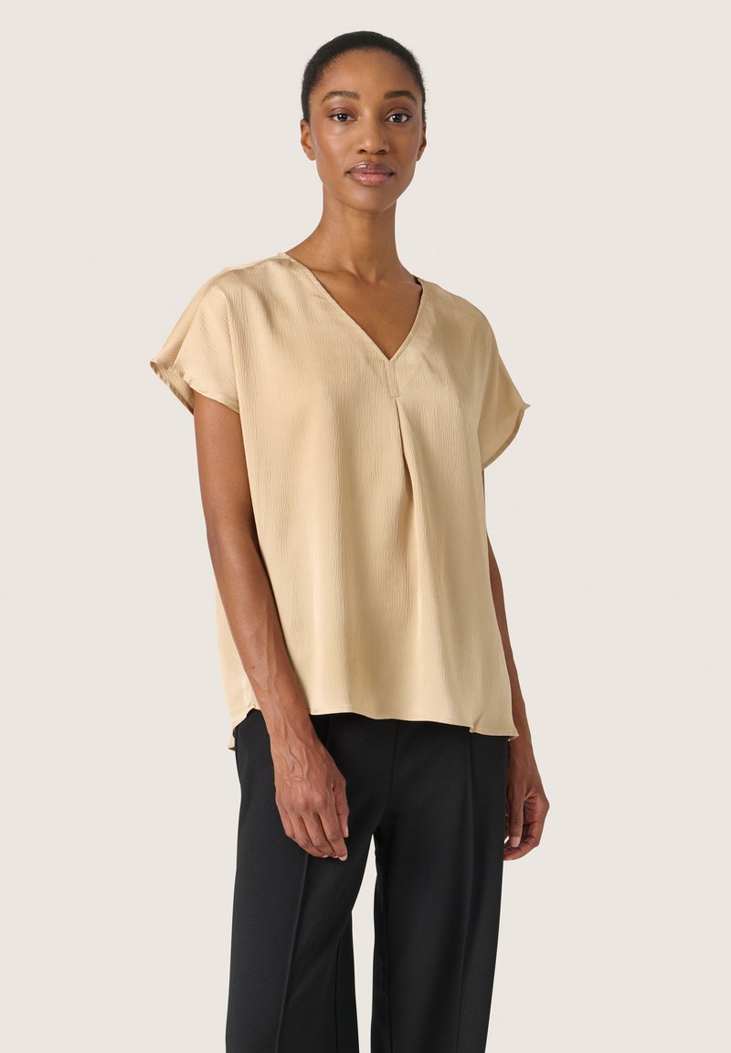 Kvinna klädd i en beige kortärmad V-ringad blus och svarta byxor, stående mot en ljus, enfärgad bakgrund.