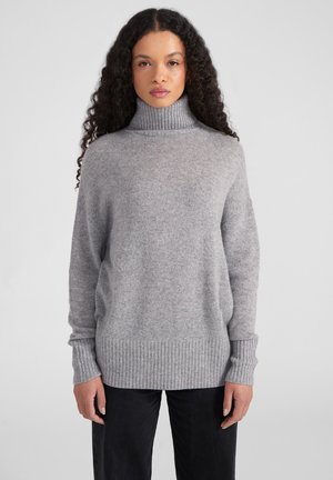 Pullover grigio a collo alto lavorato a maglia con polsini e orlo a coste, caratterizzato da una vestibilità rilassata e una texture morbida. Indossato sopra pantaloni scuri.