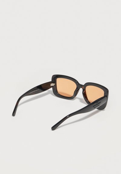 Γυαλιά ηλίου με μαύρο τετράγωνο πλαίσιο και φακούς με κεχριμπαρένιο χρώμα, ανοιχτά επάνω σε λευκή επιφάνεια.