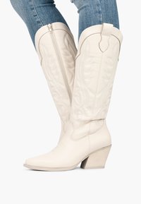 Botas de cowboy de cuero blanco con costuras decorativas, puntera ligeramente afilada y tacón apilado. Cierre de cremallera en el costado.