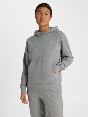 Femme portant un sweat à capuche gris et un pantalon de jogging assorti avec de petits logos en forme de chevrons, debout, une main dans la poche, sur un fond uni.