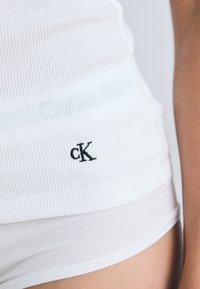 Balts ribots topiņš ar mazu melnu "cK" logo sānos, izgatavots no mīksta, elastīga materiāla, lai nodrošinātu pieguļošu izskatu.