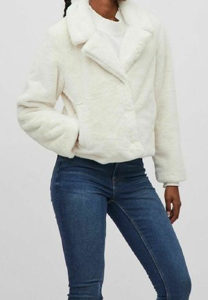 Woman wearing a white faux fur jacket over a white sweater, paired with fitted blue jeans, standing with one hand in pocket.