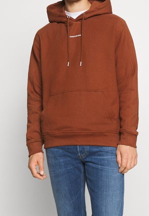 Hoodie en coton marron avec une poche frontale, des cordons de serrage ajustables et un logo blanc. Accompagné d'un jean en denim bleu.