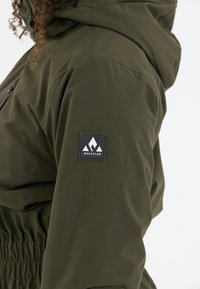 Veste imperméable verte avec taille cintrée, capuche ajustable et un patch logo noir sur le haut du bras avec des accents de design montagne.
