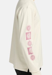 Camicia a maniche lunghe beige con motivi di sole rosa sulla manica sinistra, in cotone, taglio ampio e spacchi laterali all'orlo.