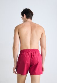 Boxeri de înot roșii cu talie elastică, având o textură netedă și o croială clasică, prezentați dintr-o vedere din spate pe un fundal neutru.