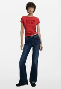 T-shirt rossa con stampa "LOVE DSGI", abbinata a jeans svasati blu scuro. Outfit completato con scarpe nere con accenti turchesi.