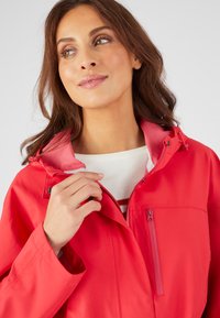 Giacca impermeabile rossa con colletto alto, tasca sul petto con zip e cappuccio regolabile con cordino. Texture liscia e design moderno.