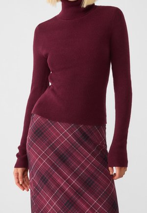 Une femme porte un pull ajusté à col roulé bordeaux à manches longues et une jupe taille haute à carreaux bordeaux, sur un fond uni.