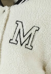 Giacca in pile bianca con una prominente lettera "M" nera ricamata sul davanti. Tessuto texturizzato con un aspetto morbido e peloso.