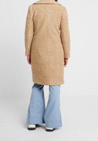 Cappotto tan, con texture, dotato di colletto e maniche lunghe. Indossato sopra pantaloni blu a palazzo. Il cappotto ha una lunghezza fino al ginocchio ed è realizzato in un tessuto morbido e peloso.