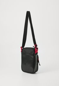 Borsa a tracolla nera con pannello frontale lucido, retro in tessuto nero e dettagli in hardware rosso. Presenta una tracolla regolabile e una chiusura con zip.