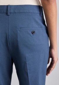 Pantaloni blu su misura con una texture liscia, dotati di una tasca posteriore con un bottone nero e una fascia in vita piatta.