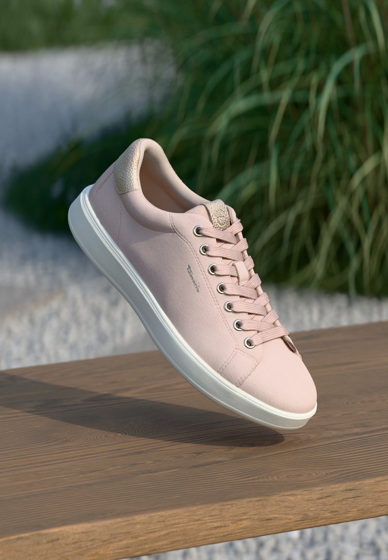 Baskets rose clair avec une semelle blanche et un détail de talon beige reposant sur une surface en bois en extérieur, avec une végétation en arrière-plan.