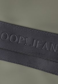 Črn tkaninski etiket z vtisnjenim besedilom "LOOP! JEANS" na olivno zeleni podlagi. Ploska struktura tkane tkanine z čistimi robovi in brez dodatnih okrasnih elementov.