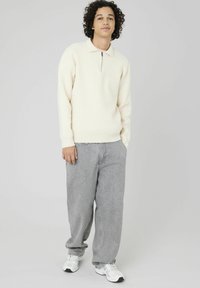 Pull en tricot crème clair avec col et zip quart, associé à un pantalon en denim gris ample et des baskets blanches. Design minimaliste, texture douce.