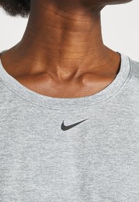 Camiseta atlética de manga corta en gris claro con escote redondo. Presenta un pequeño logo negro de Nike en el pecho. Tejido suave y texturizado.