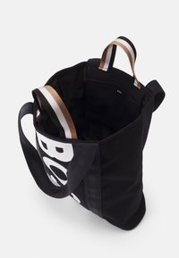 Svart tygkasse med breda svarta och gyllene randiga handtag. Har en stor vit tryckt logotyp. Insidan är fodrad med enfärgat svart tyg.