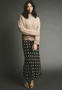 Beige Kabelstrickpullover mit geripptem Saum, kombiniert mit einem langen schwarzen Rock mit Punkten. Das Model trägt olivgrüne Stiefeletten.