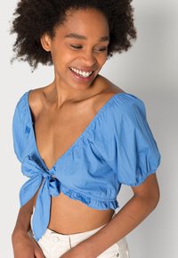 Blusa cropped em algodão de azul claro, com mangas bufantes, detalhe de laço na frente e barra franzida, criando um ajuste descontraído.