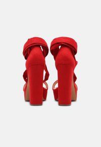 Sandales à talons hauts rouges avec une finition en daim, design à enroulement autour de la cheville et talons épais. Dos et sections des orteils ouverts pour un look audacieux.