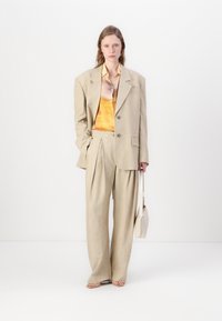 Μπέζ κοστούμι σε προσαρμοσμένη γραμμή με oversized σακάκι και πλισέ παντελόνι σε φαρδιά γραμμή, συνδυασμένο με πουκάμισο σε σχέδιο πορτοκαλί και κίτρινου χρώματος, συμπληρωμένο από μια ελαφριά τσάντα χειρός.